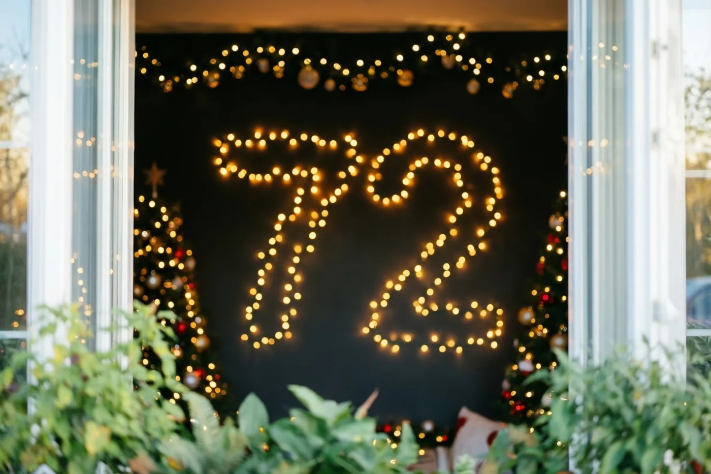 Door een raam met groen op de voorgrond zijn lichtjes in de vorm van 72 op een donkere muur te zien, met kerstbomen op de achtergrond.
