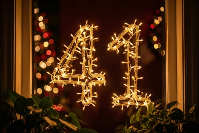 Door een raam met groene planten onderaan zijn warme lichtjes te zien die 41 vormen op een donkere muur met onscherpe feestlichtjes erachter.