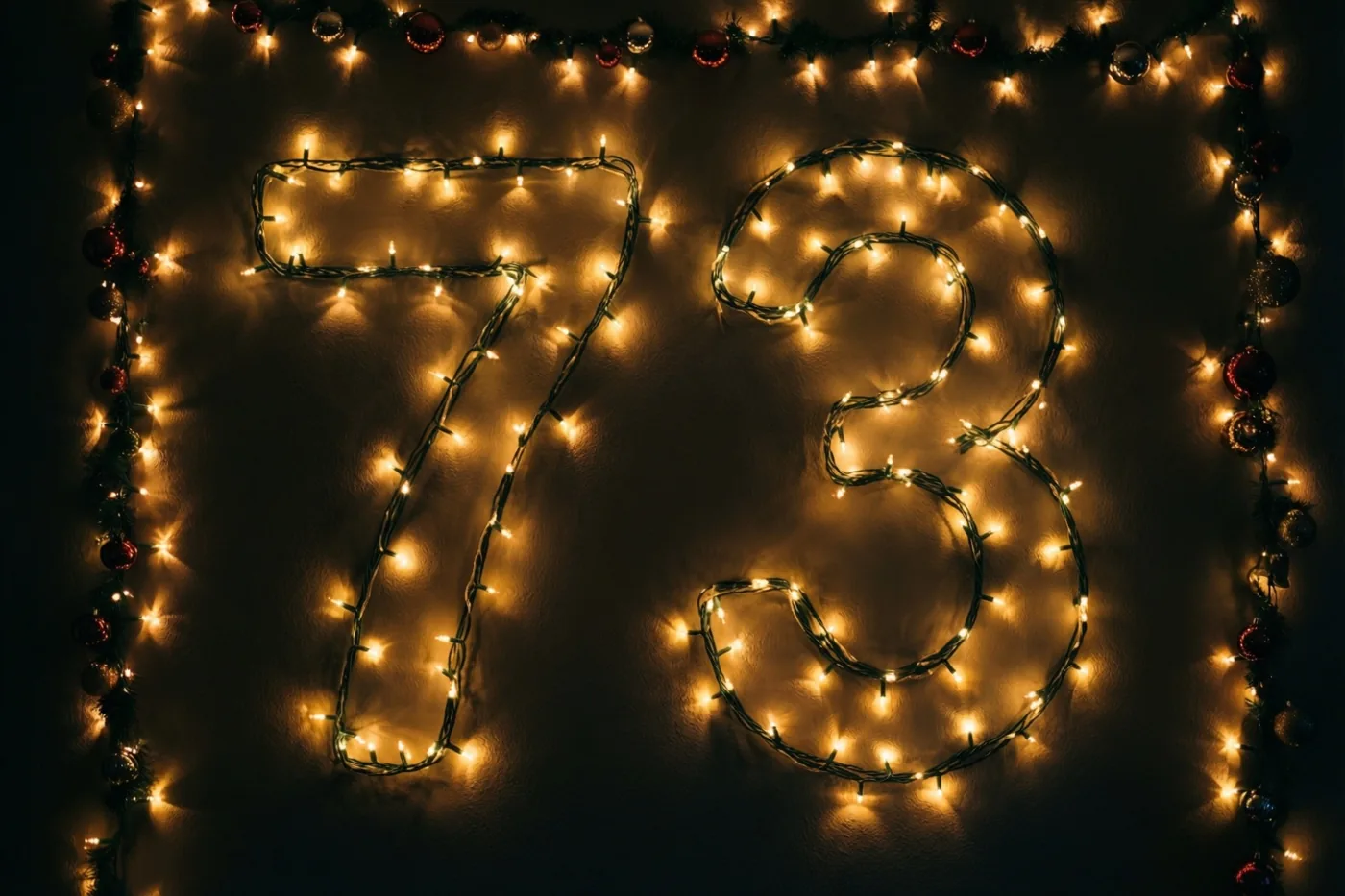 Warme lichtjes vormen 73 op een donkere muur, omlijst door een decoratieve slinger met ornamenten.