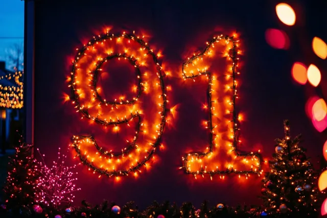 Warme lichtjes vormen 91 tegen een donkere muur, met onscherpe lichtpunten rondom en kleine versierde boompjes onderaan.