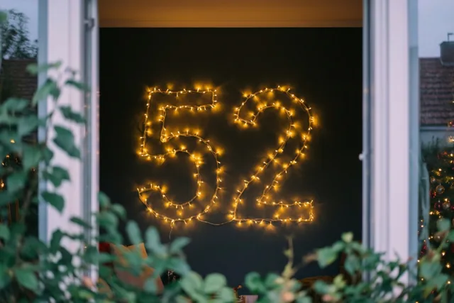 Door een open raam is het getal 52 van warme lichtjes op een donkere muur te zien, met groen op de voorgrond.