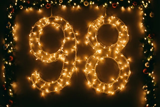 Warme lichtslingers vormen 98 tegen een donkere muur, omlijst door een versierde slinger met donkergroene en rode accenten.