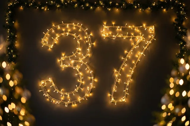 Het getal 37 van warme lichtjes hangt tegen een donkere muur, omringd door een groene slinger met verlichting en onscherpe lichtjes op de voorgrond.