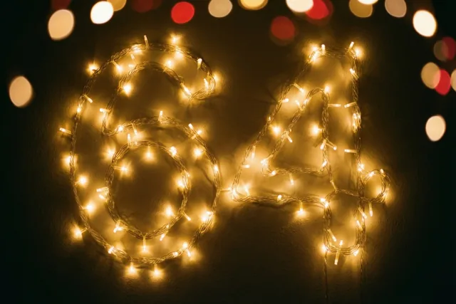 Warme lichtslingers vormen het getal 64 op een donkere muur, met kleurrijke onscherpe lichtjes op de achtergrond.