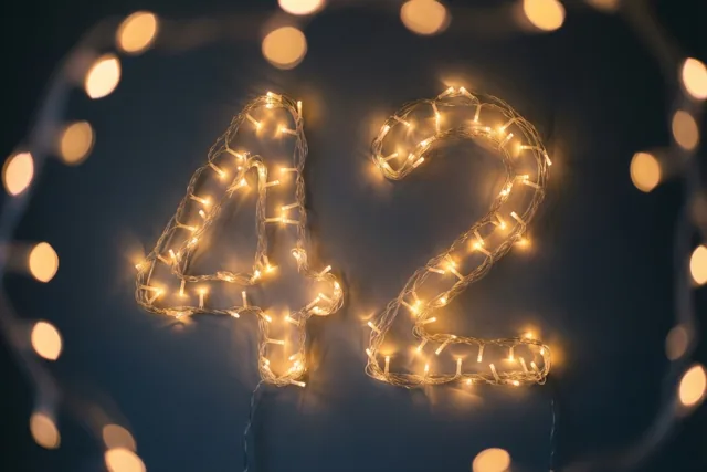 Warme lichtjes vormen 42 tegen een donkere muur, met onscherpe feestverlichting op de achtergrond.