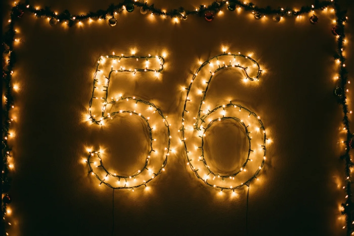 Warmgele lichtjes vormen 56 op een donkere muur, omringd door een decoratieve slinger met ornamenten.
