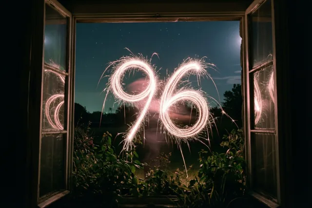 Door een open raam zijn gouden lichtsporen in de vorm van 96 te zien tegen een diepblauwe sterrenhemel, met donker gebladerte onderaan.