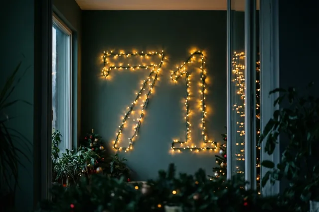 Warme lichtjes vormen 71 op een donkere blauwgroene muur, omringd door feestelijk groen en onscherpe lichtpunten.