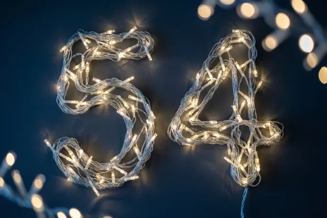Warme lichtjes vormen het getal 54 op een donkerblauwe muur, met zachte lichtvlekken op de achtergrond.