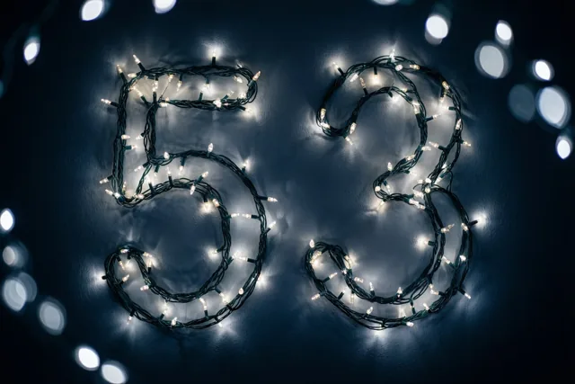 Lichtjes vormen het getal 53 op een donkere muur, met warme onscherpe lichtvlekken op de achtergrond.