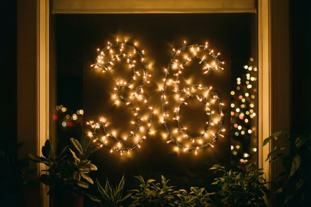 Warme lichtslingers vormen 36 op een donkere ruit, omlijst door een houten raamkozijn en groene planten, met onscherpe lichtjes op de achtergrond.