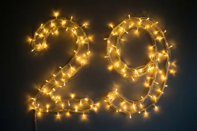 Het getal 29 is gevormd met warme lichtjes tegen een donkere muur, met zachte onscherpe lichtvlekken op de achtergrond.