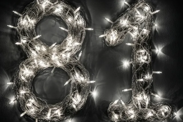 Het getal 81 is gevormd met brandende lichtslingers tegen een donkere muur, met zacht onscherpe lichtjes eromheen.