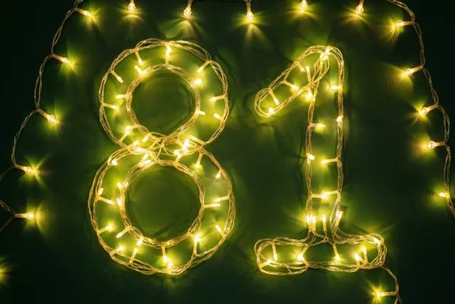 Warme lichtslingers vormen het getal 81 tegen een donkergroene muur, met zacht onscherpe lichtjes in beeld.