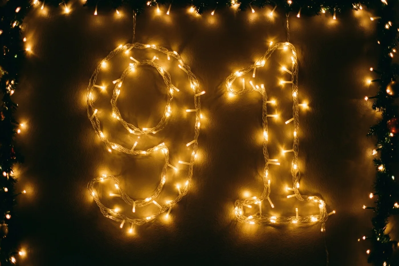 Warme lichtjes vormen 91 tegen een donkere muur, omkaderd door decoratief groen en zachte goudgele lichtvlekken.