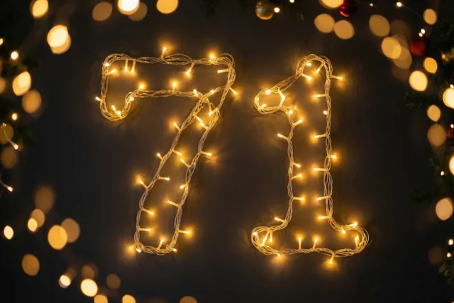 Warme lichtjes vormen 71 op een donkere muur, omringd door dennentakken en onscherpe feestlichtjes.