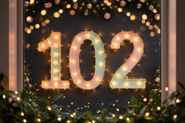 Warme lichtslingers vormen 102 op een donkere muur, omringd door feestelijk groen en onscherpe lichtjes in zachte tinten.