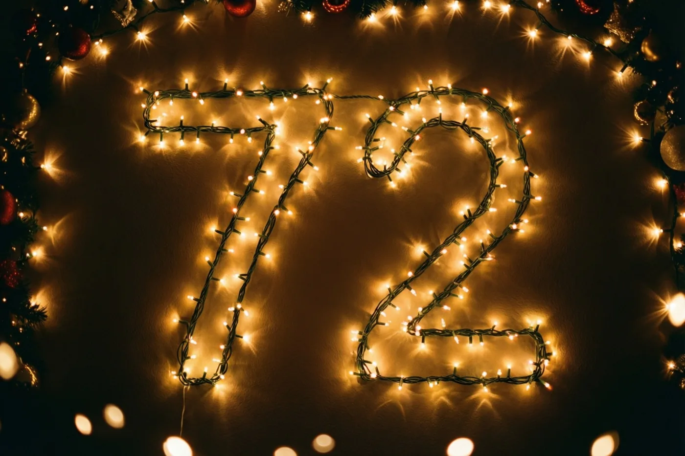 Warme lichtjes vormen 72 op een donkere muur, omringd door onscherpe lichtpunten en feestversiering.