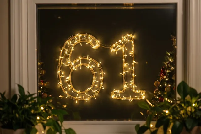 Warme lichtjes vormen 61 tegen een donkere muur, omlijst door een wit raam en geflankeerd door groen en kleine kerstbomen.