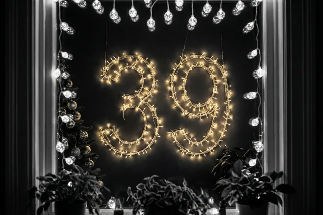 Warme lichtjes vormen 39 op een donkere muur, omringd door decoratieve verlichting en planten in een verder zwart-wit beeld.