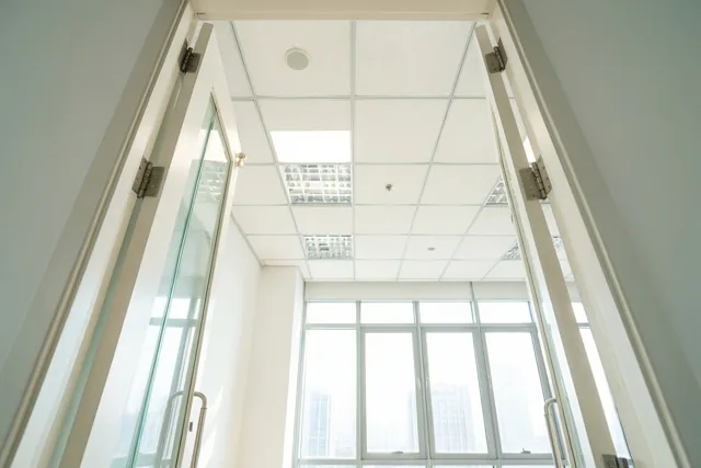 Een open deuropening geeft zicht op een lege, lichte kantoorruimte met een hoog plafond en grote ramen in witte en lichtgrijze tinten.