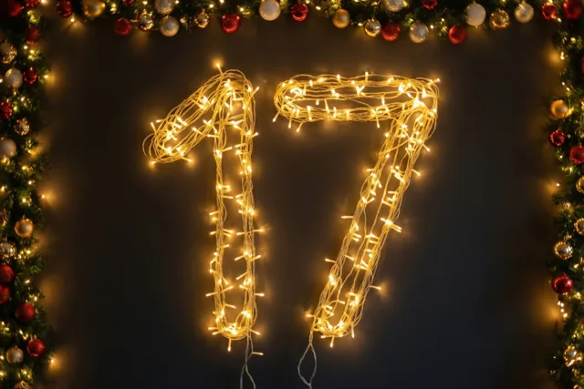 Warme lichtjes vormen 17 tegen een donkere muur, omlijst door een decoratieve slinger met rode en gouden versieringen.