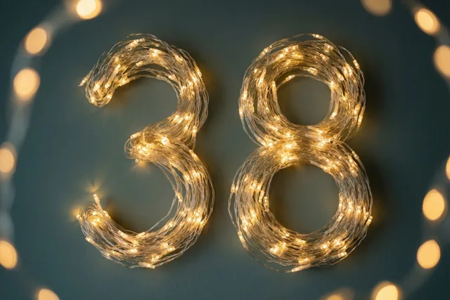 Warme lichtjes vormen het getal 38 tegen een donkere muur met onscherpe lichtvlekken op de achtergrond.