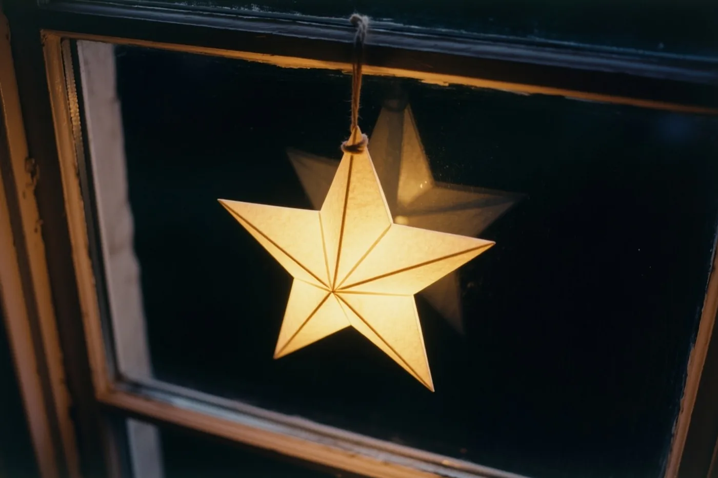 Een lichtgevende papieren ster hangt aan een touwtje in een donker raam met een houten kader, met een lichte rand rond de foto.