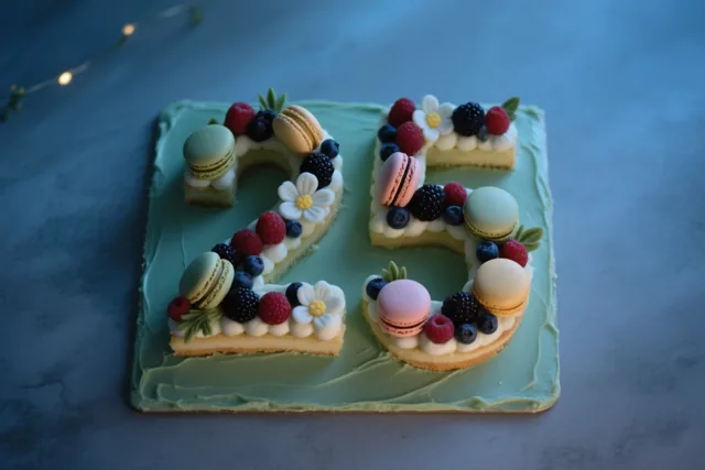 Een cijfertaart in de vorm van 25 met lichtgroen glazuur, bessen, macarons en eetbare bloemen staat tegen een blauwe achtergrond met linksboven zachte lichtslingers.