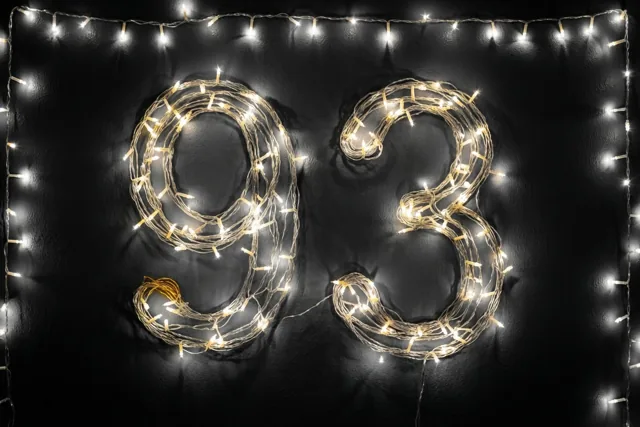 Warme lichtjes vormen 93 op een donkere muur met structuur, omringd door een rand van extra lichtjes.