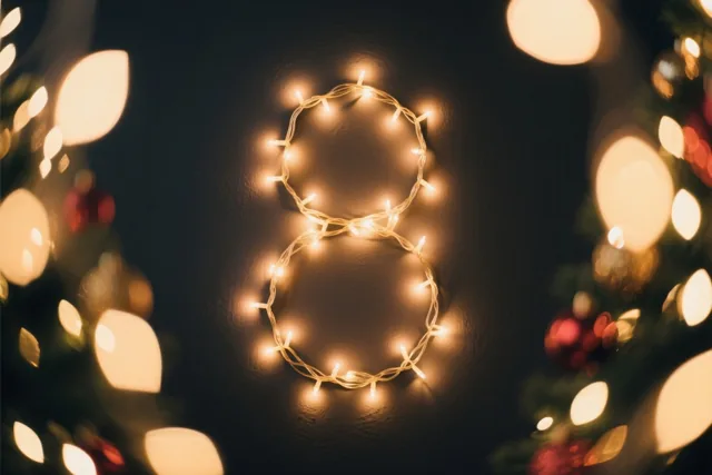 Een 8 van warme lichtjes hangt tegen een donkere muur, omringd door onscherpe kerstlichtjes en versieringen in gouden en rode tinten.