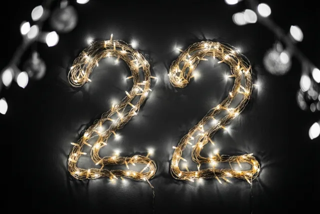 Het getal 22 is gemaakt van warme lichtjes tegen een donkere muur met wazige feestverlichting op de achtergrond.