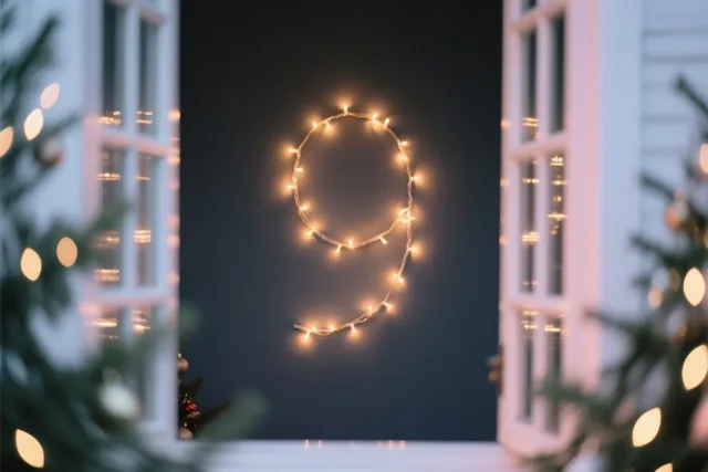 Een 9 van warme lichtjes hangt op een donkere muur, omkaderd door witte raamruiten en onscherpe lichtjes van een kerstboom in gouden en donkerblauwe tinten.