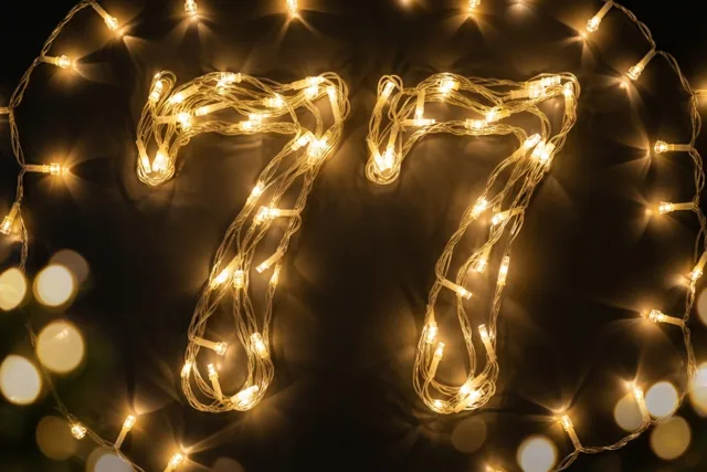 Het getal 77 is gevormd uit warm gloeiende lichtslingers tegen een donkere muur, met onscherpe lichtjes op de achtergrond.