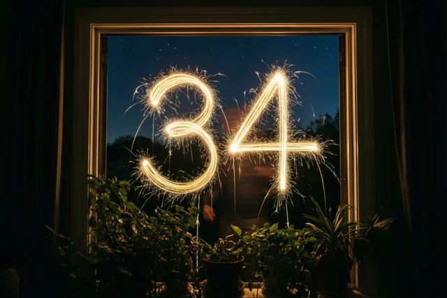Door een raam zijn gouden lichtsporen in de vorm van 34 tegen een sterrenhemel te zien, met donkere gordijnen en potplanten op de voorgrond.