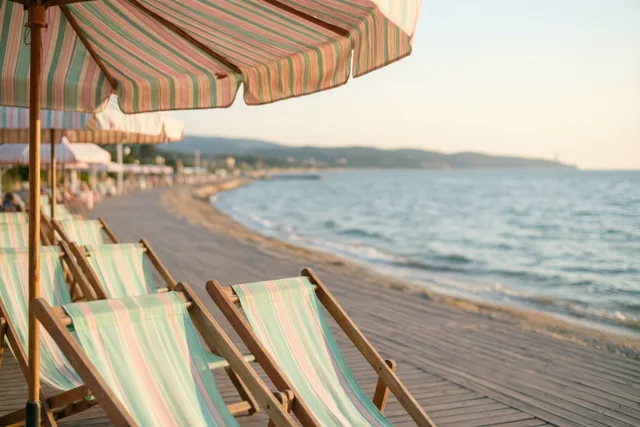 Een rij gestreepte ligstoelen met bijpassende zonneschermen staat op een houten promenade aan een kalme zee met een heiige horizon onder een zachtgekleurde, warme lucht.