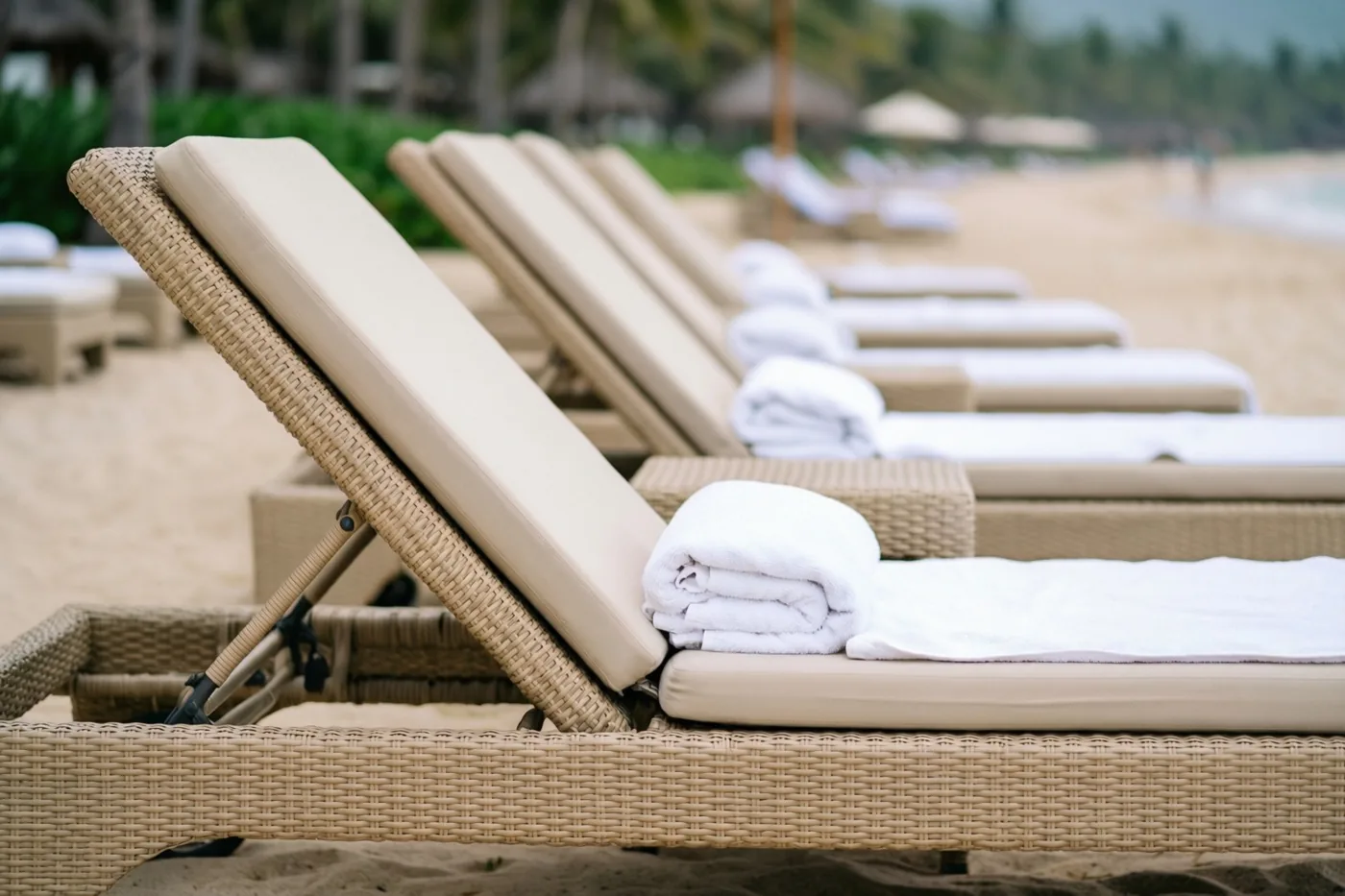 Rijen beige rotan ligstoelen met witte handdoeken staan op een zandstrand, met onscherpe palmbomen en de zee op de achtergrond.