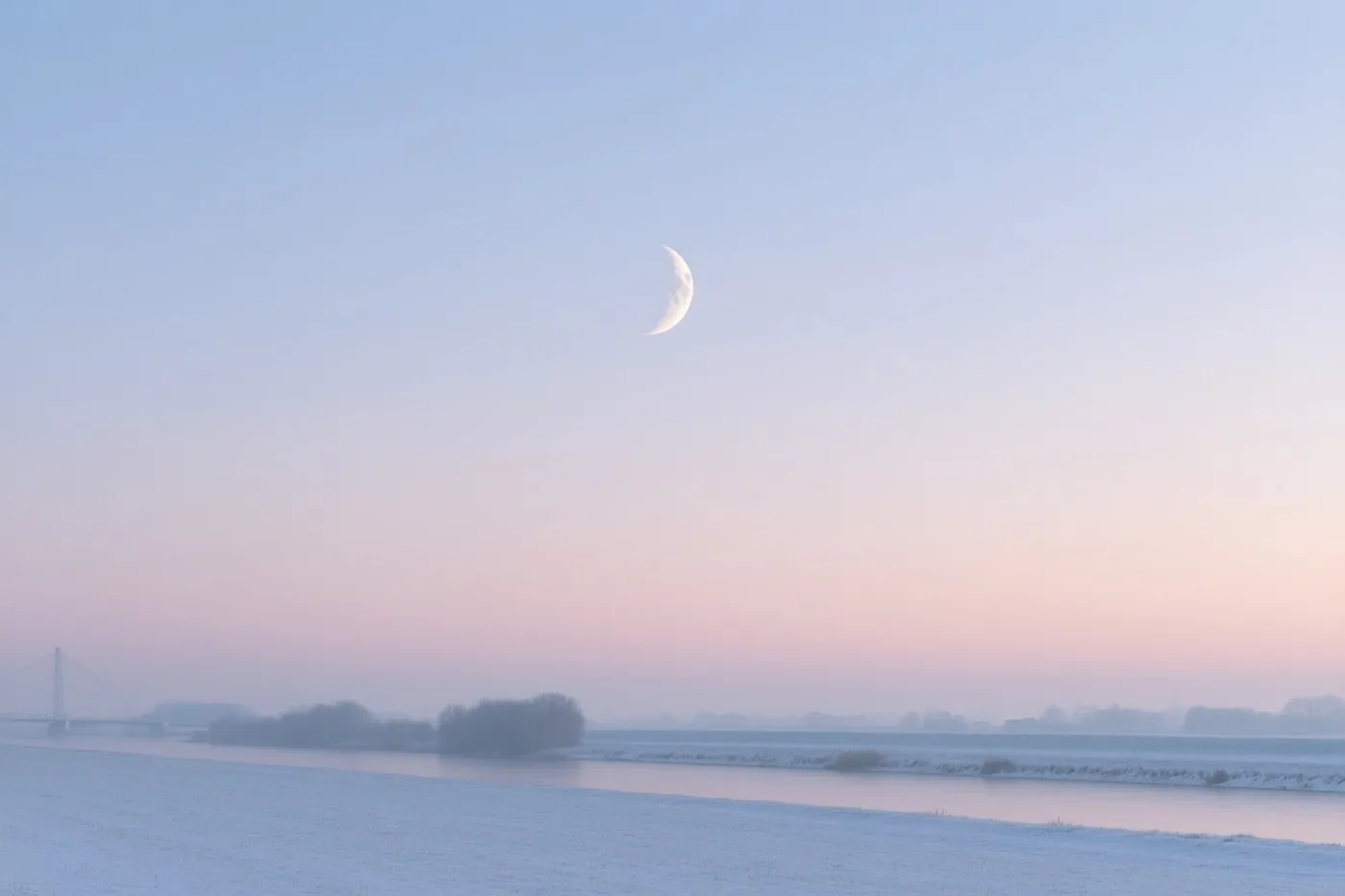 Een dunne maansikkel staat boven een bevroren rivier, met een verre brug en kale bomen in de mist onder een winterhemel in blauw en roze.