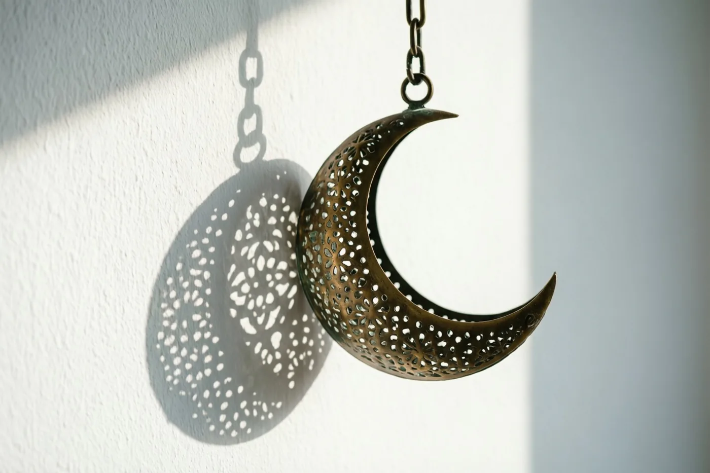 Een messing maansikkellantaarn met fijne opengewerkte patronen hangt aan een ketting en werpt in natuurlijk zonlicht een gedetailleerde schaduw op een witte muur.