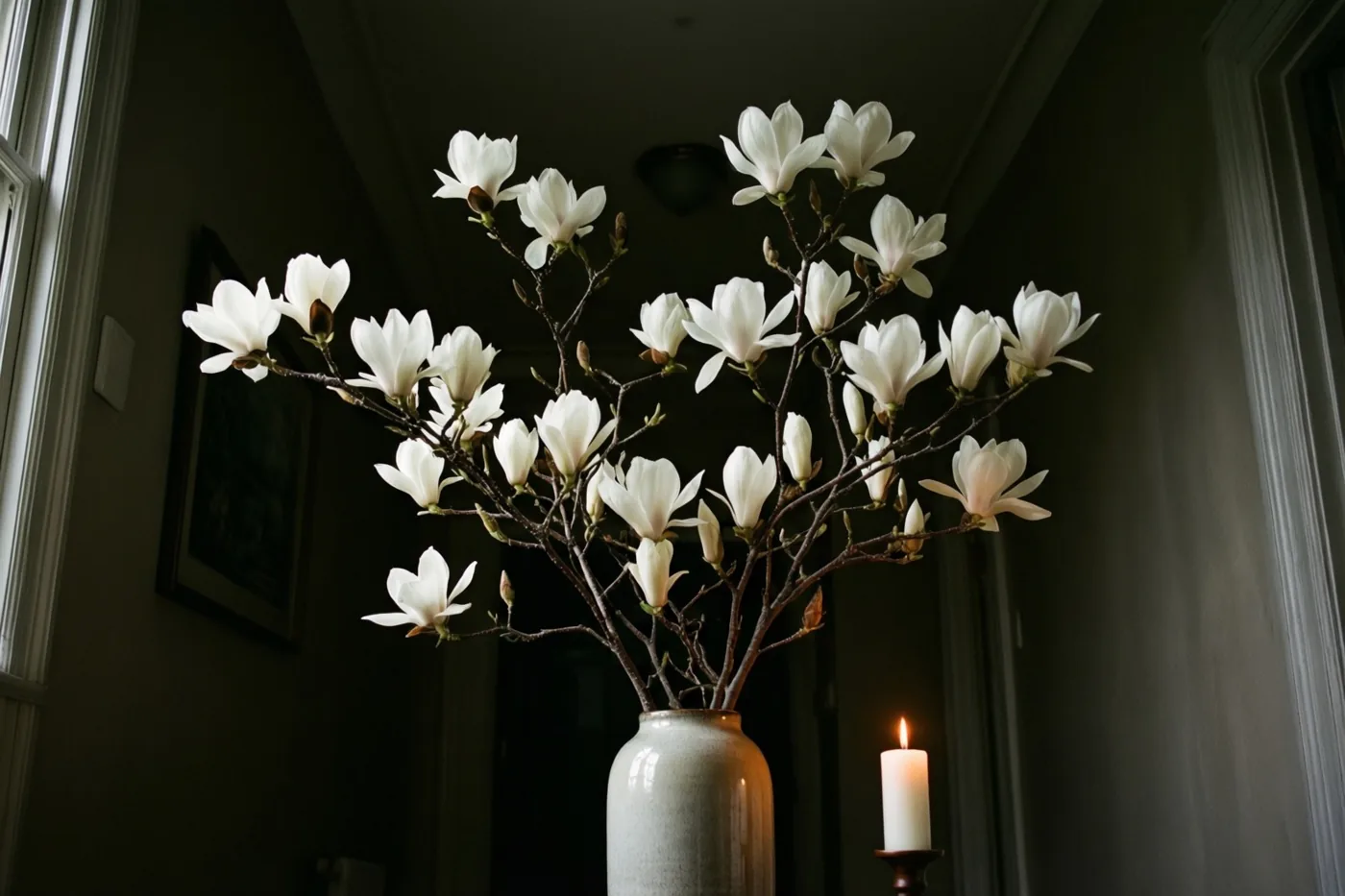 Een witte magnoliatak staat in een hoge keramieken vaas in een schemerige gang, verlicht door daglicht uit een raam en een brandende kaars op een standaard ernaast.