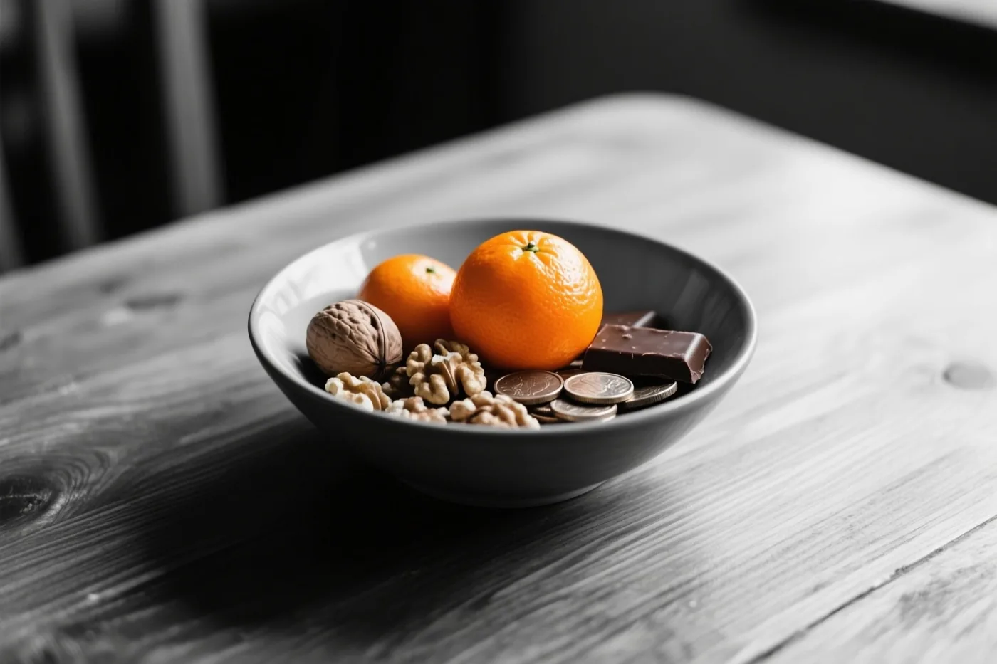 Een donkere kom met mandarijnen, walnoten en stukken chocolade staat op een houten tafel, met het fruit en de chocolade in kleur tegen een monochrome achtergrond.