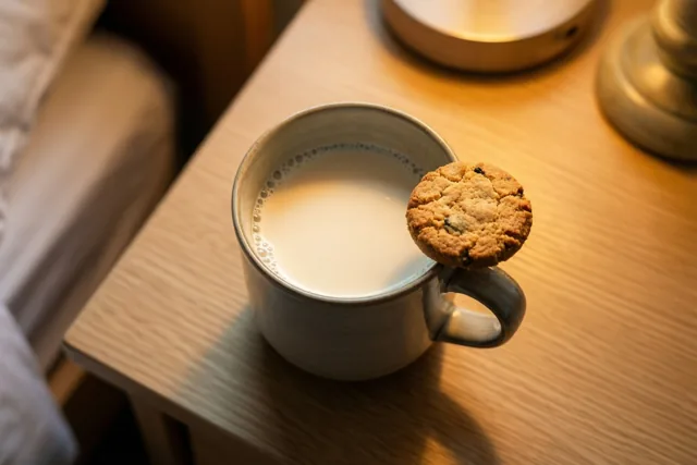 Een kleine keramieken mok melk met een chocoladekoekje op het oor staat op een houten nachtkastje in warm ochtendlicht.