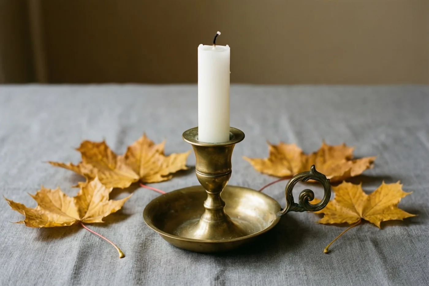 Een hoge witte kaars in een messing kandelaar staat op grijs linnen tussen gouden herfstbladeren, tegen een neutrale achtergrond.