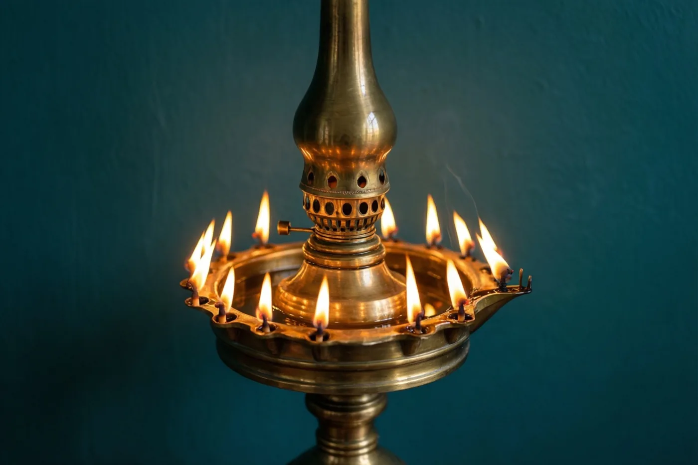 Een hoge messing olielamp met meerdere brandende lonten staat tegen een donkere blauwgroene achtergrond.