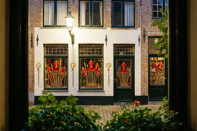 Door een donkere omlijsting is een Vlaamse straat bij nacht te zien, met rode mijters en gouden staven in warm verlichte winkelramen tussen bakstenen en witgekalkte muren.
