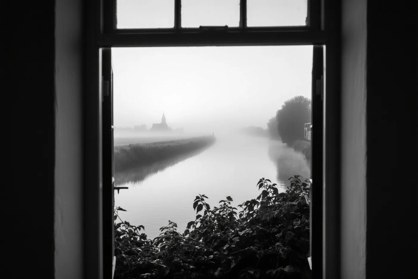 Een zwart-witbeeld door een raam toont een rustige rivier met lage ochtendmist en een kerktoren in de verte.