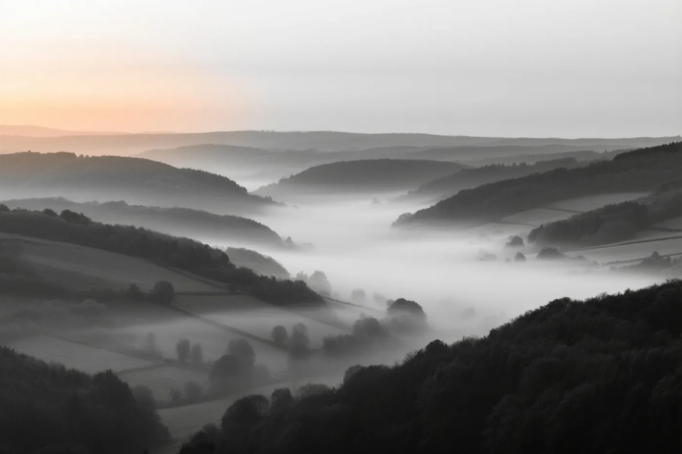 Mist bedekt delen van velden en bossen in een heuvelachtige vallei bij zonsopgang, met een oranje gloed aan de horizon.