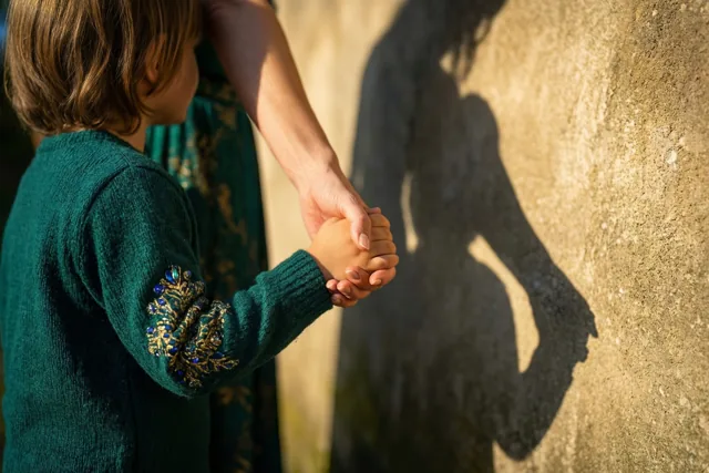 Een moeder en kind in groene kleding houden elkaars hand vast, van achteren gezien, en werpen samen één schaduw op een zonovergoten muur met structuur.