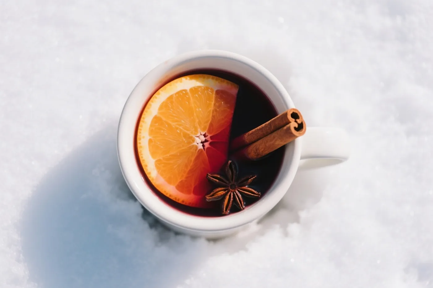 Een witte mok met donkere glühwein, een schijfje sinaasappel, een kaneelstokje en steranijs staat in verse sneeuw.