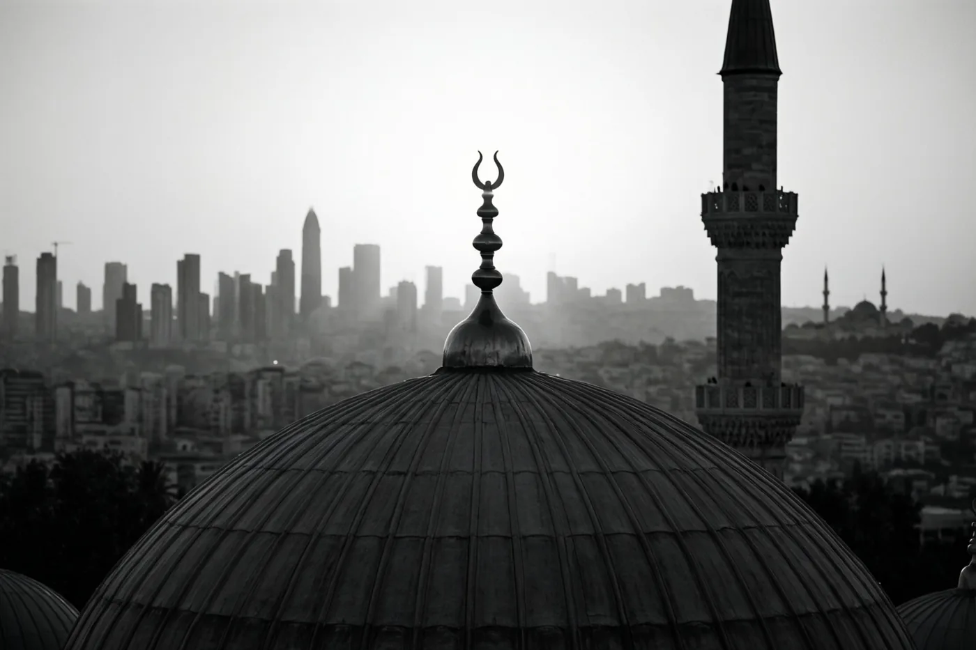 Een zwart-witfoto toont het silhouet van een moskeekoepel met een maansikkel, een minaret rechts en een wazig stadsgezicht op de achtergrond.